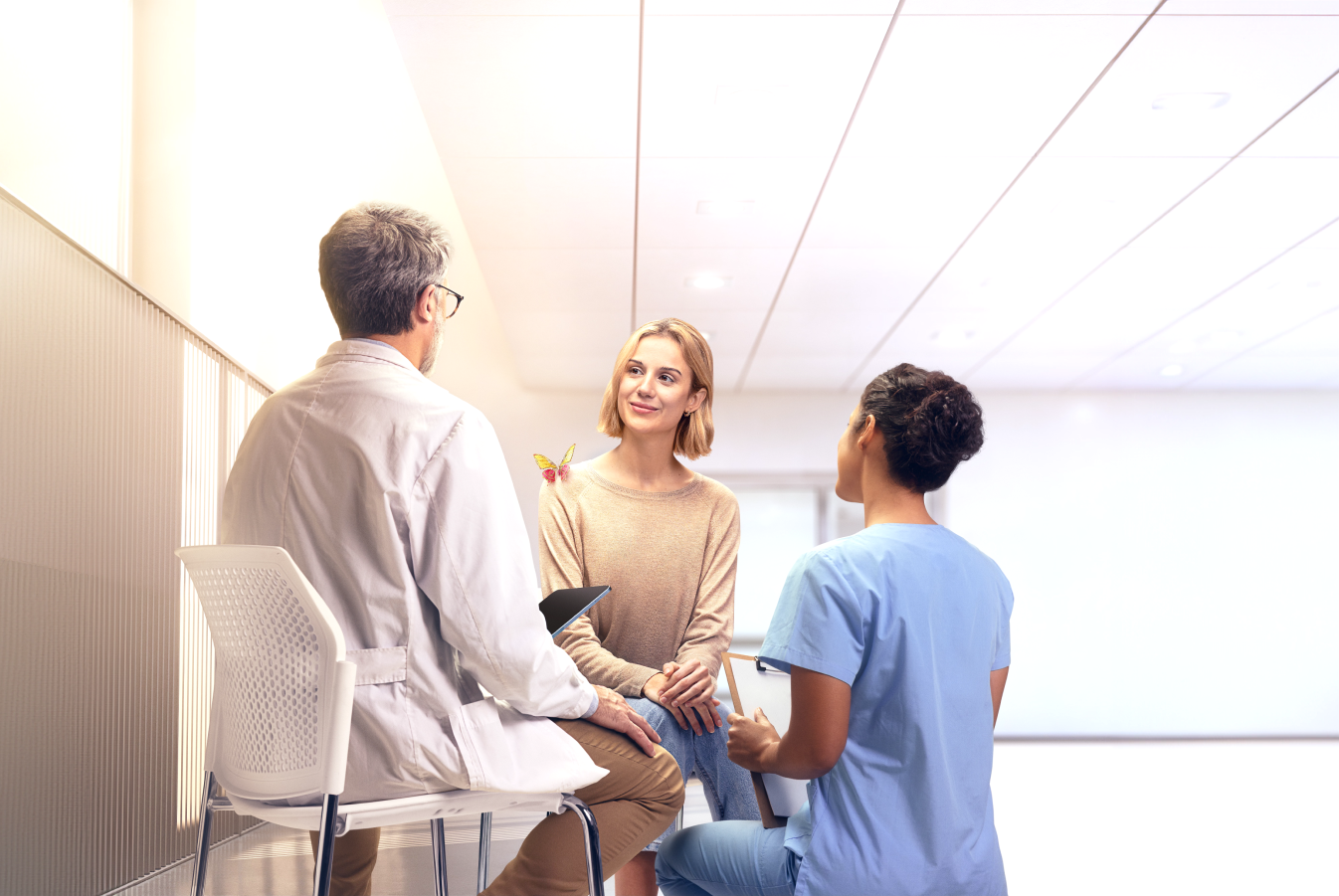 Photo image of an adult female patient speaking with doctor and nurse in a hospital setting, with an engineered mechanical yellow and magenta butterfly on her shoulder